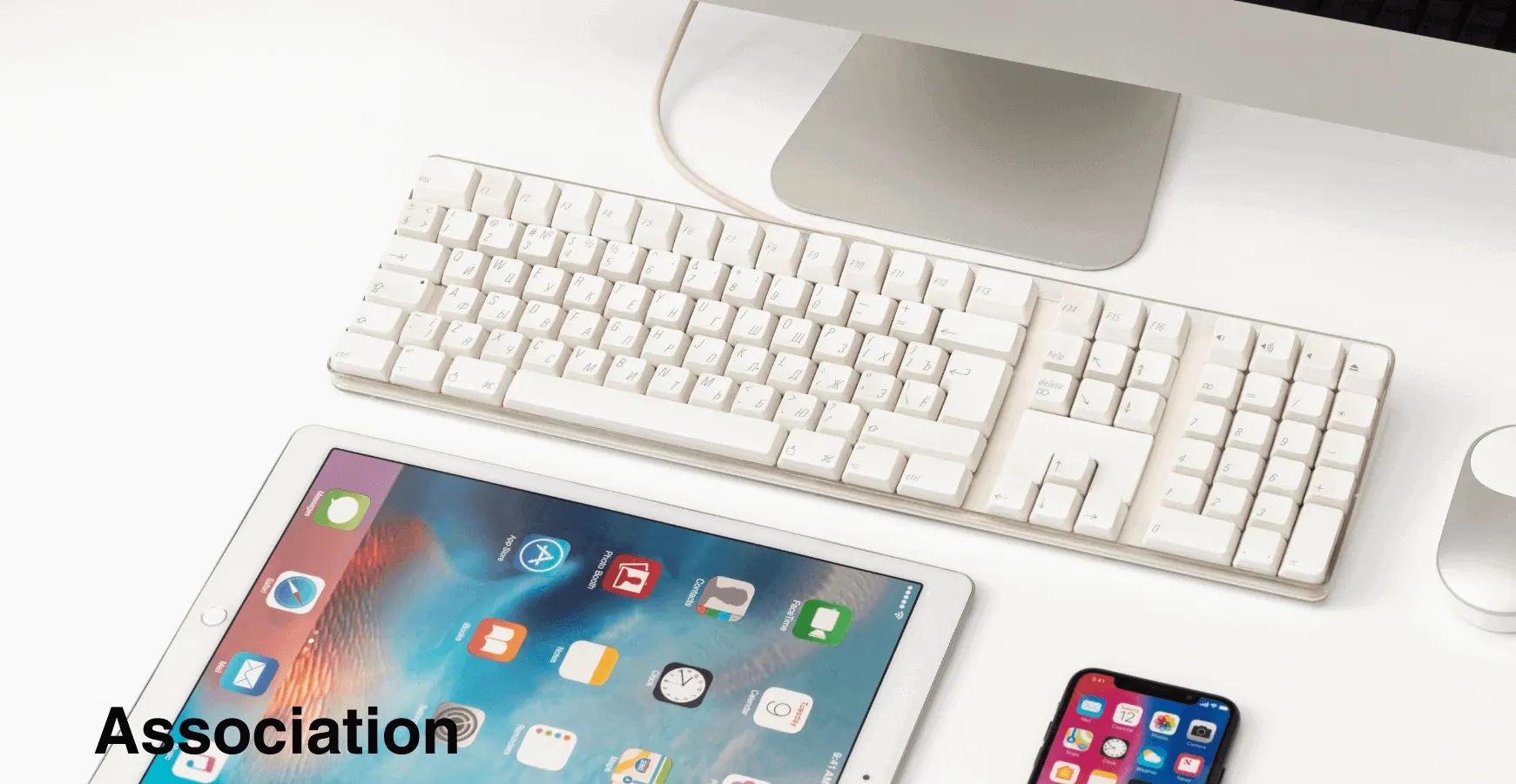 Ordinateur de bureau moderne avec clavier blanc, tablette iPad affichant des applications, et smartphone sur un bureau propre – symbolisant les outils numériques utilisés par les associations.