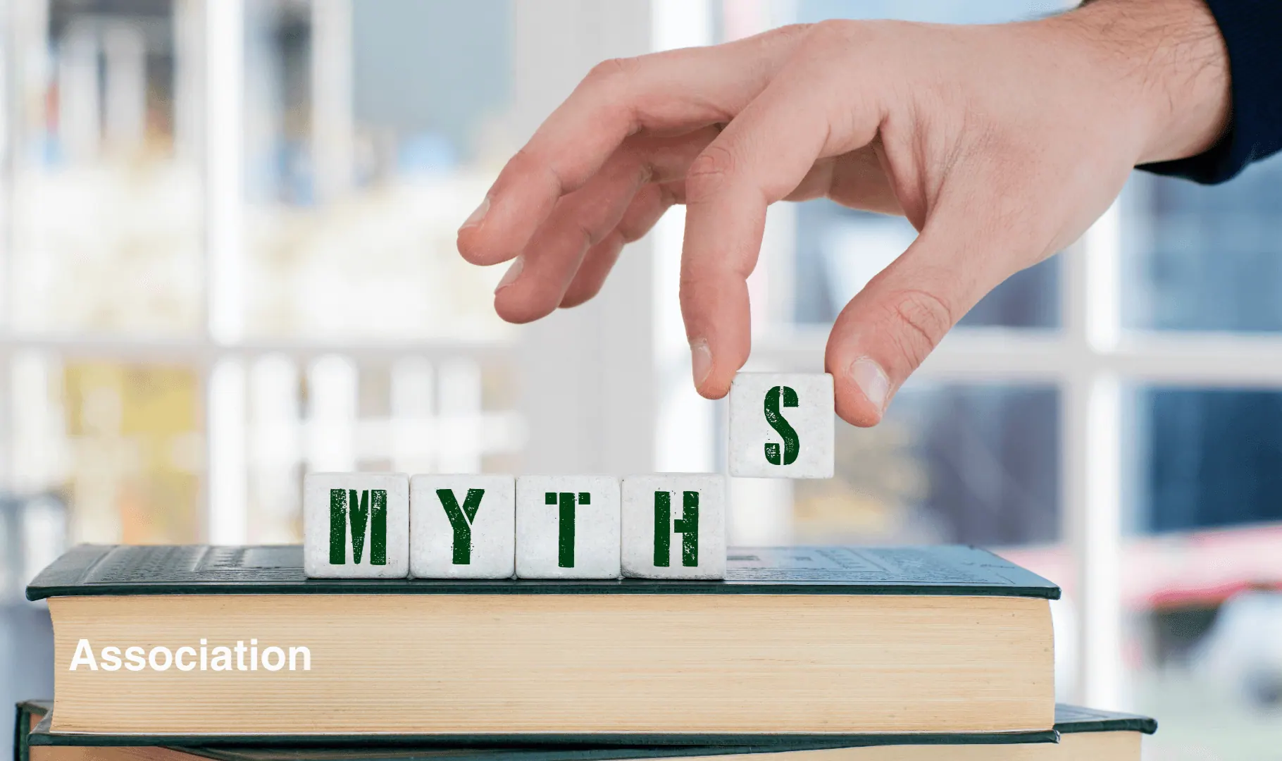 12 mythes sur Google Ad Grants. Main d’une personne manipulant des cubes formant le mot "MYTHS" posés sur un livre, illustrant la déconstruction des idées reçues sur les associations et le numérique.